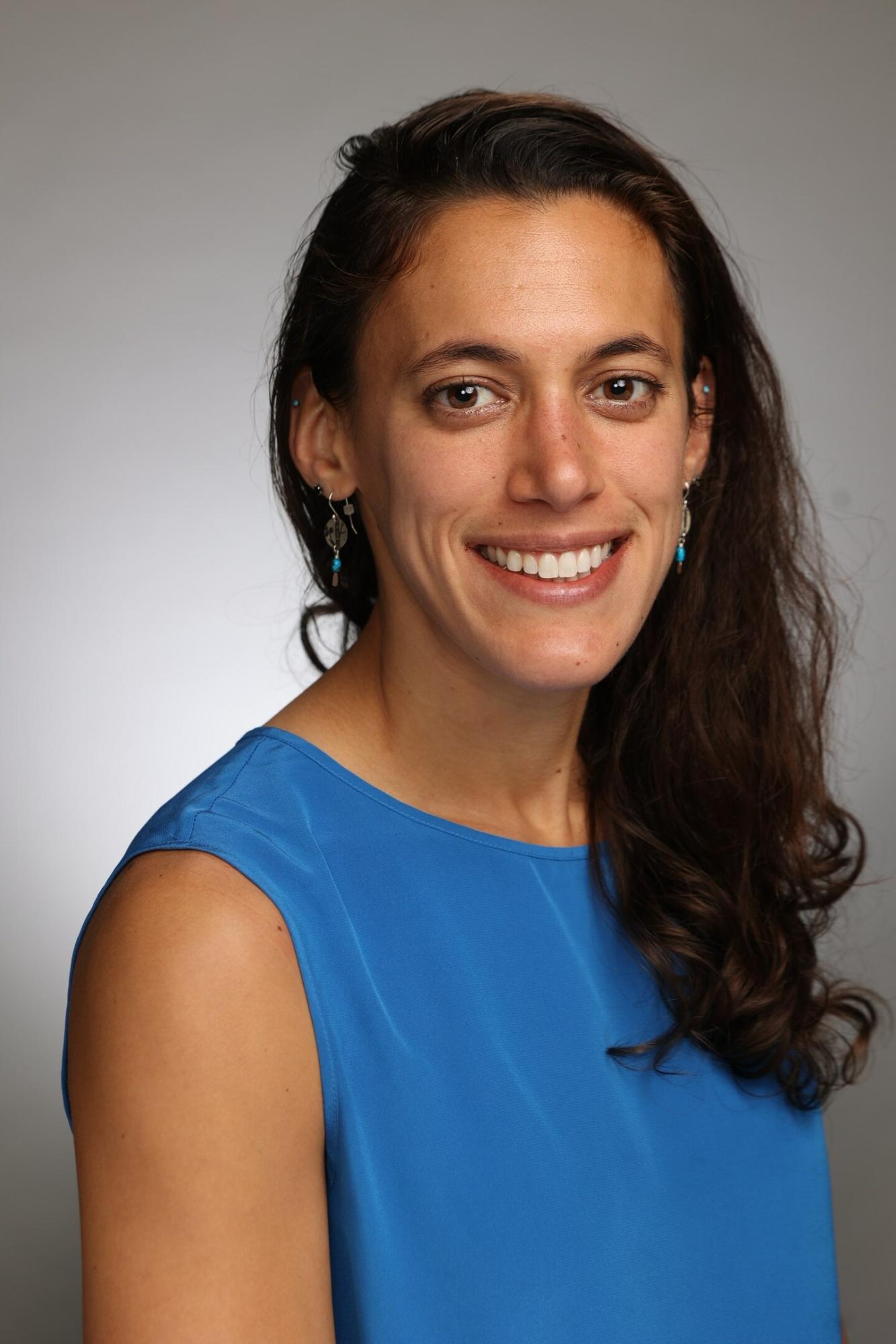 Girl with brown hair which is down smiles at camera with blue shirt.